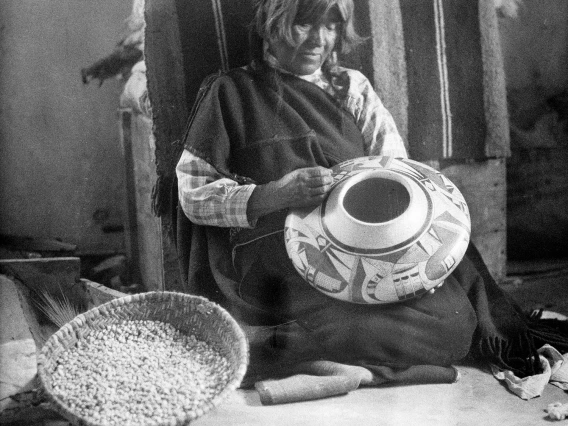 A Hopi woman sits on the ground as she appears to be paining a ceramic vessel she has made.