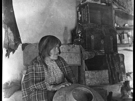 A Hopi woman with a shawl around her shoulders sits on the floor of a dwelling or store holding a large ceramic olla in front of her