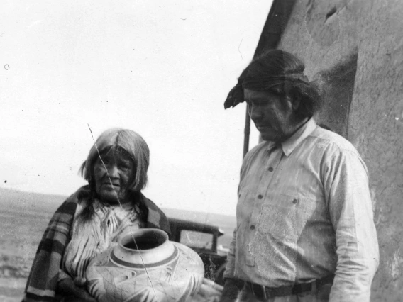 A Hopi woman and man stand outside a dwelling. The woman is hold a large pot. The man to her left is looking at the pot.