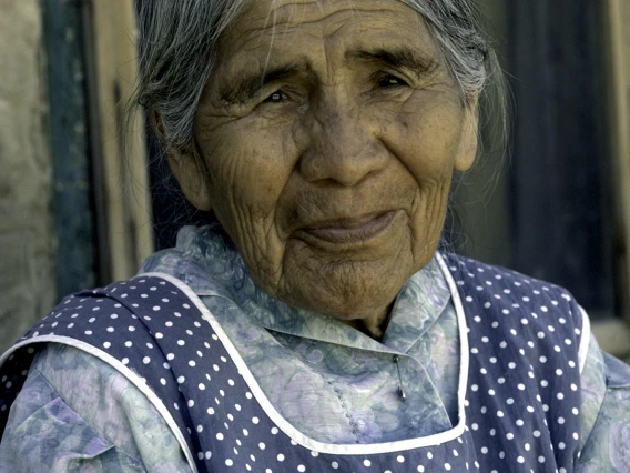 An elderly Hopi woman with short salt-and-pepper hair smiles at the camera. She is wearing a blue floral dress and over it is a blue apron with white polka dots.