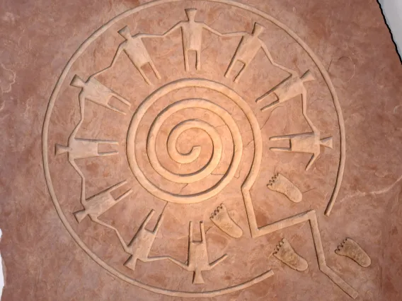 This image is of a sandstone-carved symbol illustrating the paths of life concept, with hand-holding figures standing in a circle above a spiraliform maze. The piece is by Hopi artist Gerald Dawavendewa.