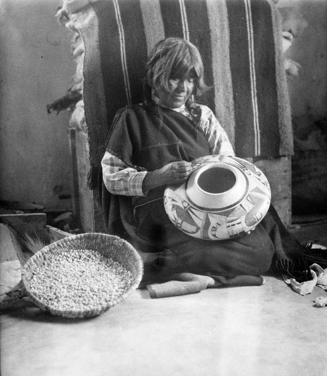 A Hopi woman sits on the ground as she appears to be paining a ceramic vessel she has made.