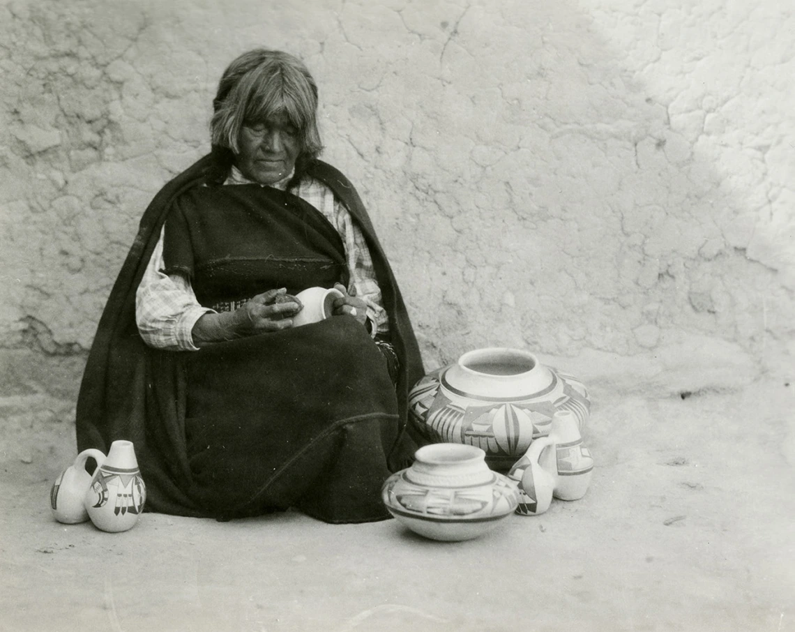 a Hopi woman sits on the ground in front of an adobe wall, sanding a small ceramic vessel she is sanding. She is surrounded by other vessels she has already made.