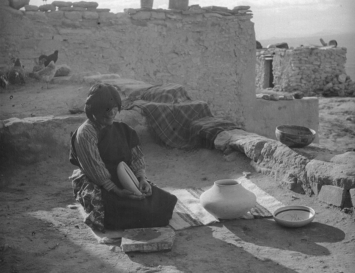 A Hopi woman sits on a woven textile which is spread on the ground. She is holding and is surrounded by pieces of pottery she has made.