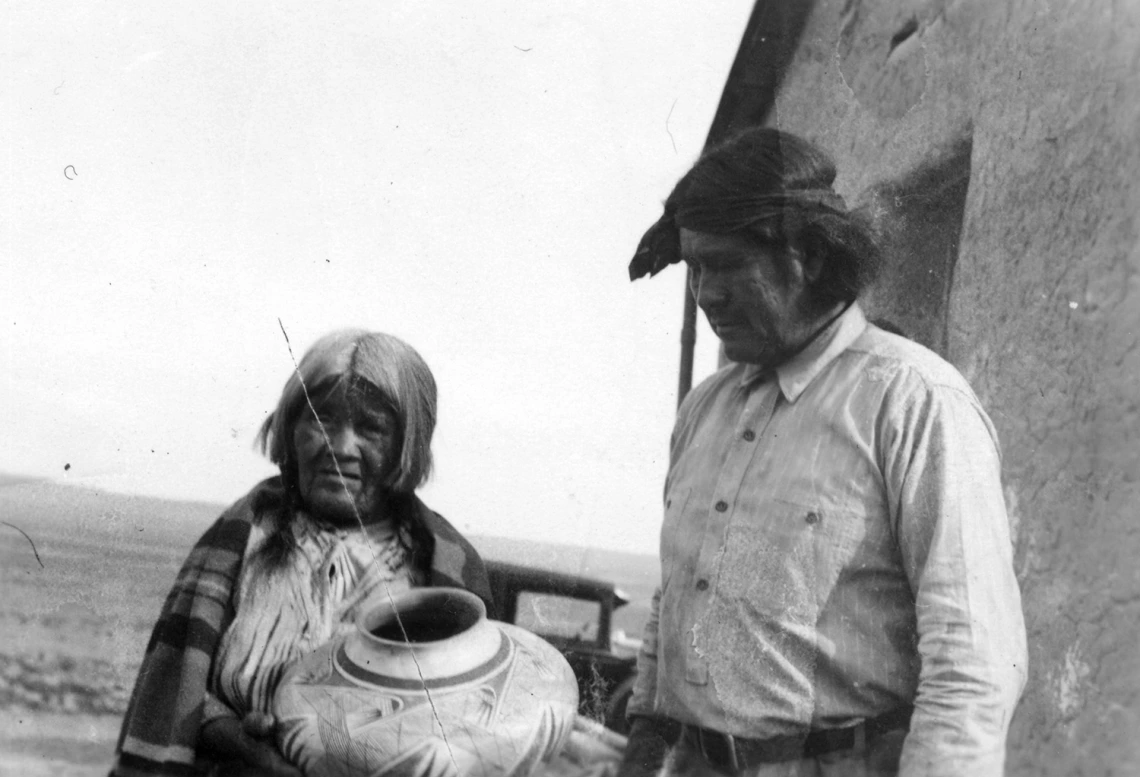 A Hopi woman and man stand outside a dwelling. The woman is hold a large pot. The man to her left is looking at the pot.