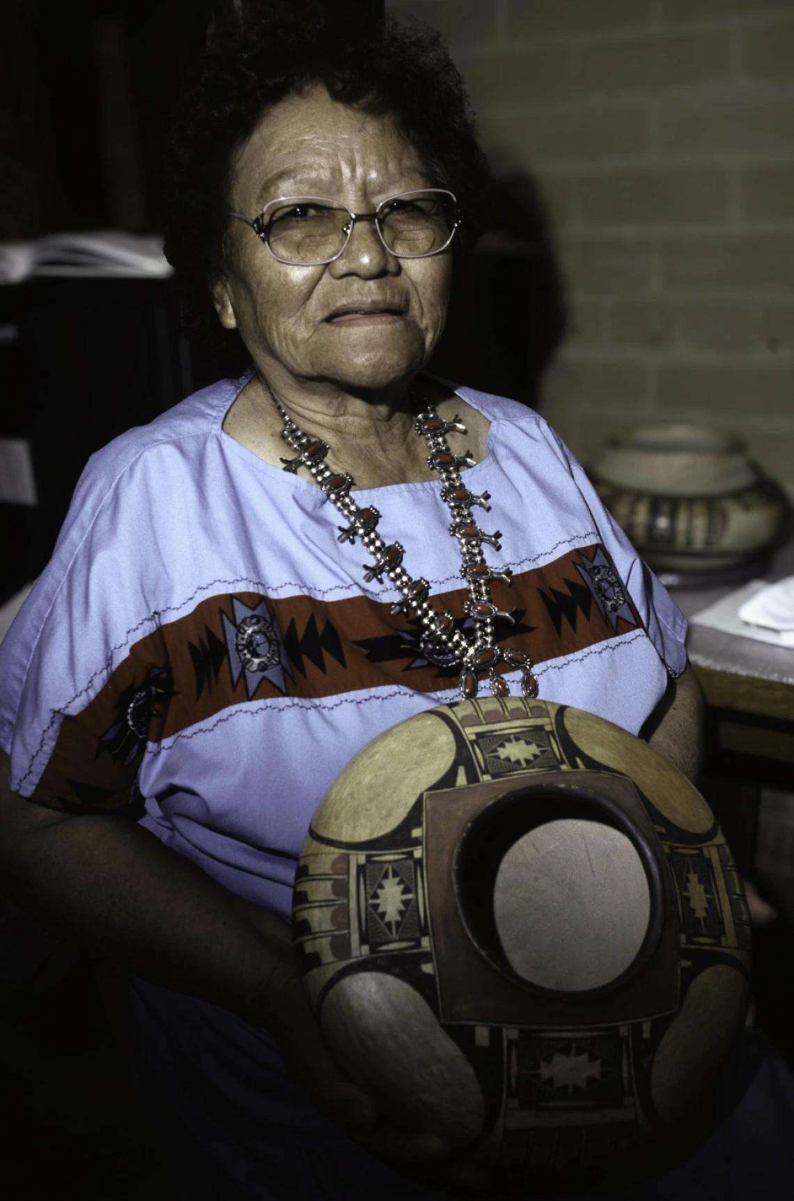 An older woman with short dark hair and wearing glasses, holds a Hopi pot in front of her. She is wearing a blue dress and a silver with coral quash blossom necklace.