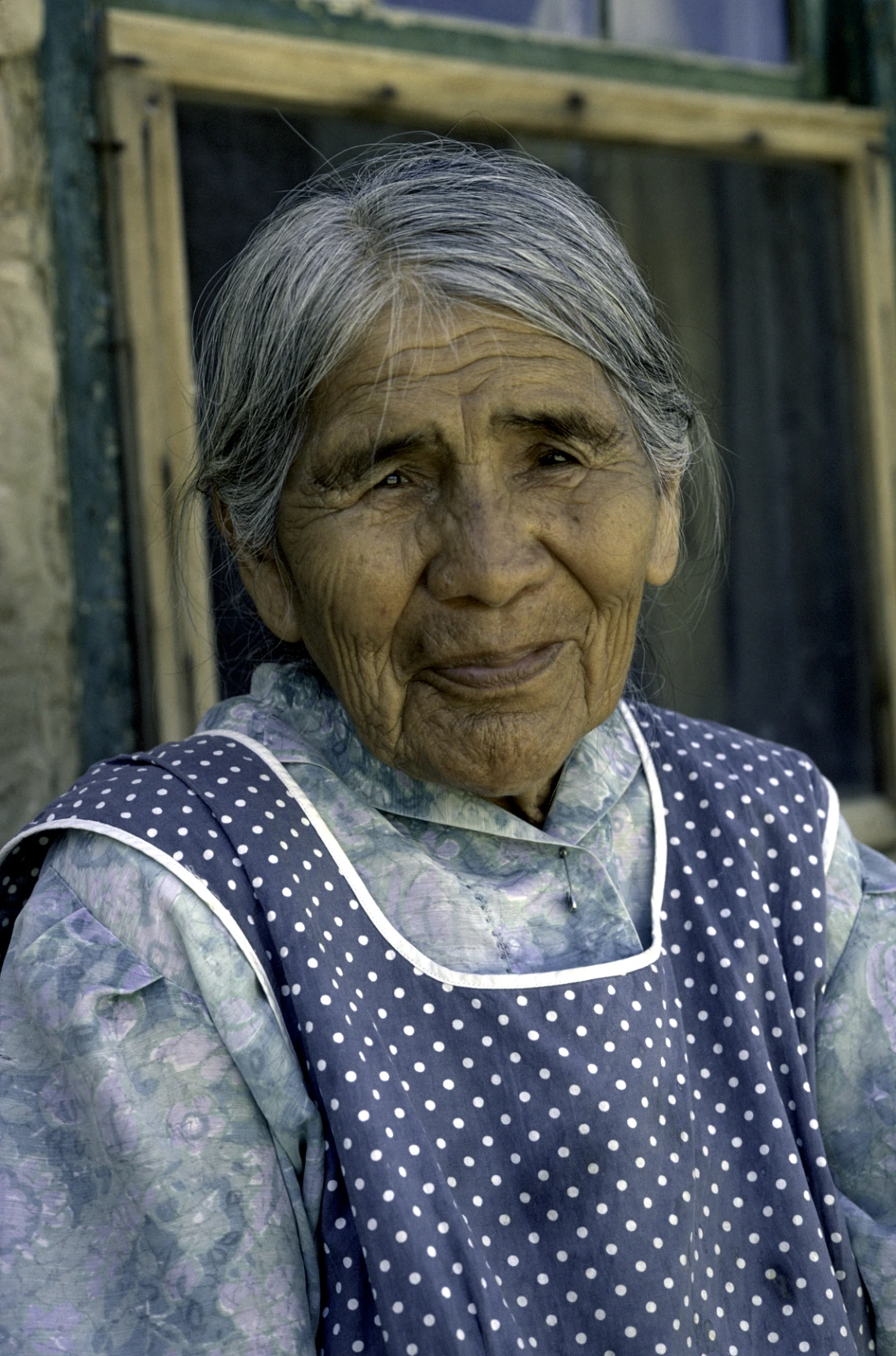 An elderly Hopi woman with short salt-and-pepper hair smiles at the camera. She is wearing a blue floral dress and over it is a blue apron with white polka dots.