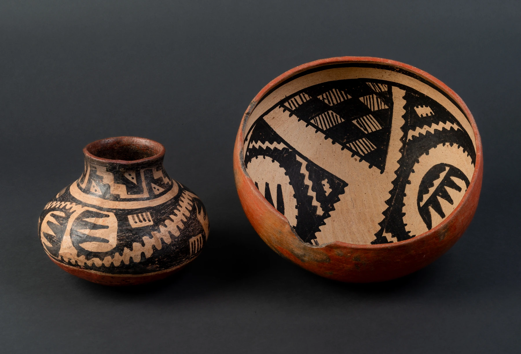 Two Gila Polychrome ceramic vessels sit side by side. On the left is a small jar. On the right is a bowl. Both have painted black designs on a white field. The ceramic fabric itself is red