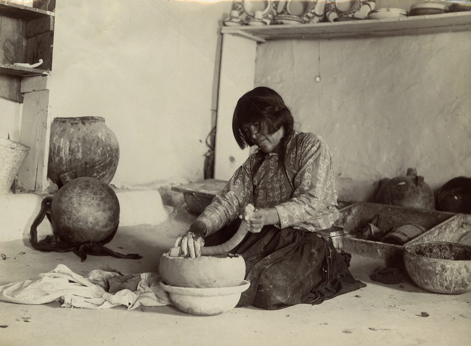 A Hopi woman sits on a dirt floor as she coils a ceramic vessel in her home studio.